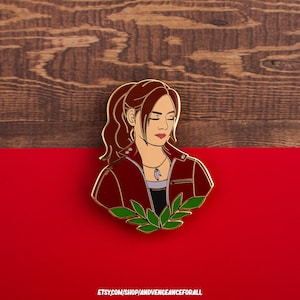 Peut inclure: Une épingle en émail dorée représentant une femme aux cheveux bruns et une veste rouge. Elle porte un collier avec un pendentif. L'épingle est ornée d'un motif de feuilles vertes en bas.  ETSY.COM/SHOP/ANDVENGEANCEFORALL