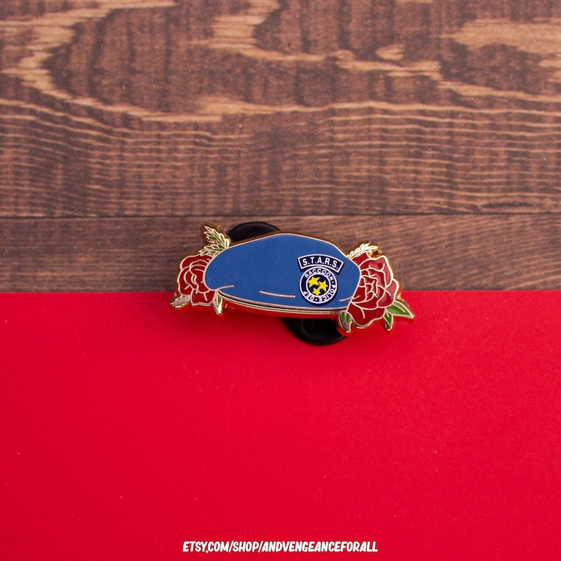 K&ouml;nnte beinhalten: Ein blauer Barett-Pin mit der Aufschrift "S.T.A.R.S. Raccoon City" und einem gelben Emblem, verziert mit roten Rosen. Auf rotem Untergrund vor einem Holzmaserungshintergrund. Die Etsy-Shop-URL ist unten sichtbar.