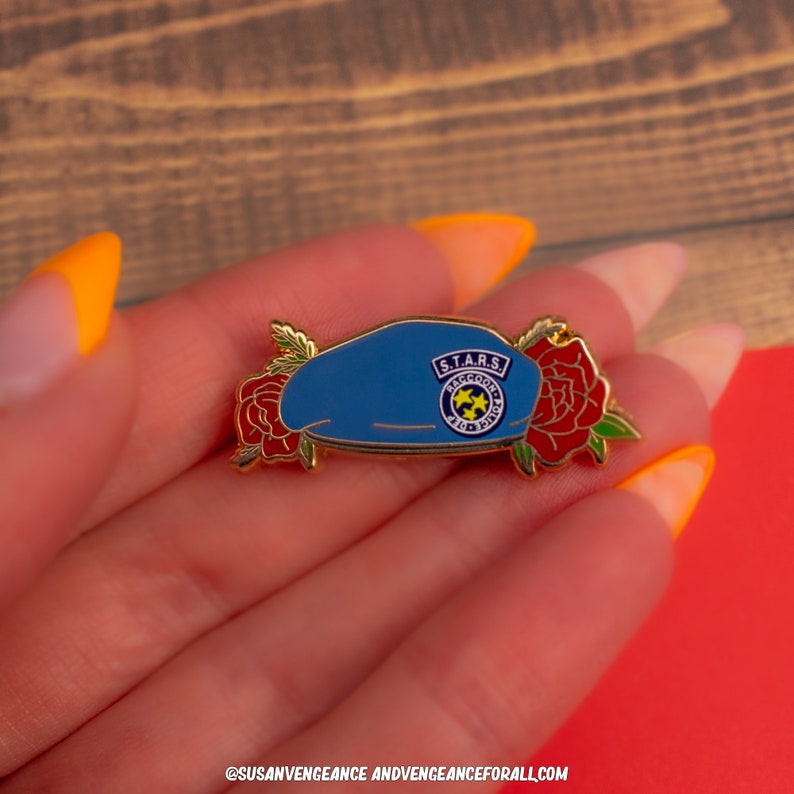 K&ouml;nnte beinhalten: Ein blauer Emaille-Pin mit einem Barett-Design mit dem Text "S.T.A.R.S. Raccoon City" und einem gelben Emblem. Der Pin ist mit roten Rosen und goldenen Akzenten verziert. Der Pin befindet sich auf einem roten und holzmaserierten Hintergrund.