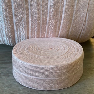 Puede incluir: Un rollo de cinta el&aacute;stica rosa p&aacute;lido est&aacute; delante de una tela a juego. La cinta est&aacute; enrollada apretadamente, mostrando c&iacute;rculos conc&eacute;ntricos. La tela detr&aacute;s tiene rayas verticales. El color es un rosa suave y neutro.