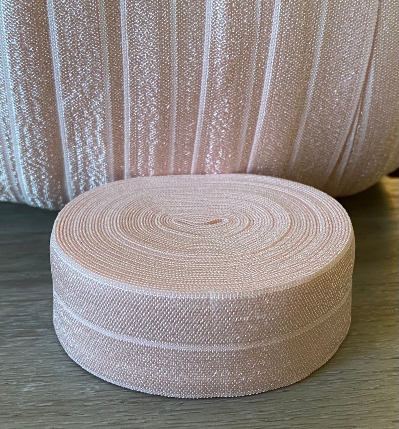 Puede incluir: Un rollo de cinta el&aacute;stica rosa p&aacute;lido est&aacute; delante de una tela a juego. La cinta est&aacute; enrollada apretadamente, mostrando c&iacute;rculos conc&eacute;ntricos. La tela detr&aacute;s tiene rayas verticales. El color es un rosa suave y neutro.