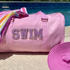 May include: A pink  swim bag with the word "SWIM" in purple letters with gold glitter accents. The bag is next to a striped towel and a pink sun hat.