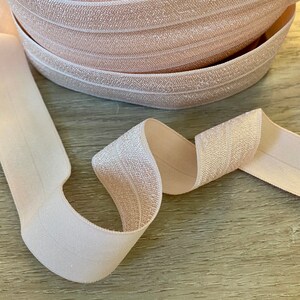 Puede incluir: Un rollo de cinta el&aacute;stica rosa p&aacute;lido con un brillo sutil. La cinta es plana y tiene una superficie texturizada con finas l&iacute;neas blancas que recorren su longitud. Una porci&oacute;n de la cinta est&aacute; desenrollada, mostrando su flexibilidad y textura.