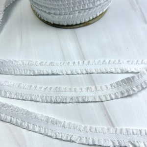 May include: White elastic trim with a ruffled edge. The trim is shown in a close-up view, with multiple layers of the trim visible.