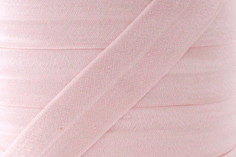Puede incluir: Primer plano de una bobina de cinta al bies rosa p&aacute;lido. La tela est&aacute; enrollada firmemente, con una tira diagonal de la cinta visible en la imagen. La textura de la tela es ligeramente visible, sugiriendo un material tejido.