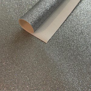 May include: A roll of silver glitter fabric with a white backing. The fabric is partially unrolled, revealing the texture and sparkle of the glitter. The background is also covered in the same silver glitter.
