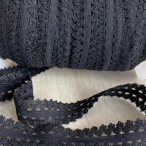 May include: A roll of black lace ribbon with a floral edge. The ribbon is made of a delicate, open-weave material. The ribbon is unrolled, showing the intricate floral design along the edge. The ribbon is on a light-colored surface.
