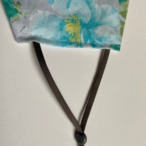 May include: A close-up of a gray fabric face mask with a floral pattern. The mask has brown adjustable straps with a black plastic slider.