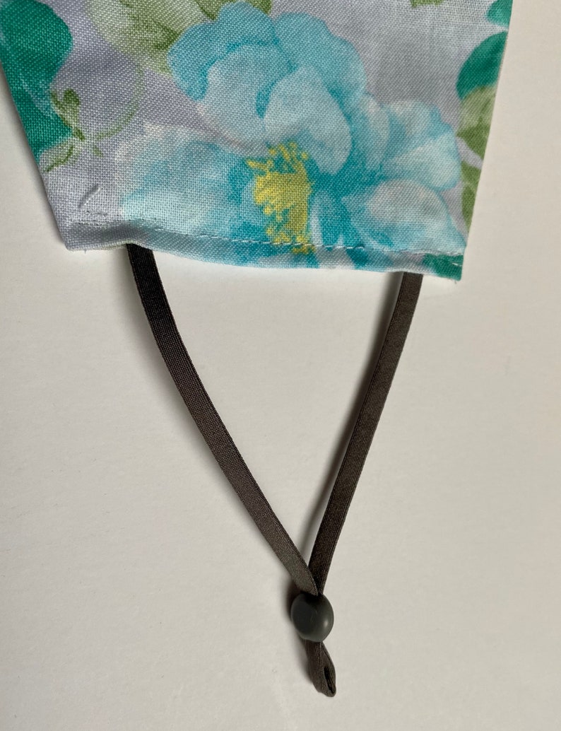 May include: A close-up of a gray fabric face mask with a floral pattern. The mask has brown adjustable straps with a black plastic slider.