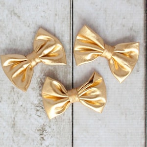 May include: Three gold metallic bow hair clips on a white wood background.