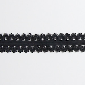 May include: Black lace trim with a floral design. The trim features a row of small, flower-shaped embellishments along the top and bottom edges, connected by a solid black band. This decorative trim is suitable for various craft projects.
