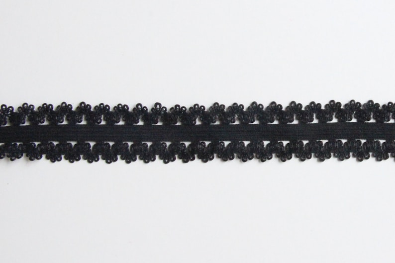 May include: Black lace trim with a floral design. The trim features a row of small, flower-shaped embellishments along the top and bottom edges, connected by a solid black band. This decorative trim is suitable for various craft projects.