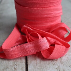 May include: A roll of coral pink elastic fold over elastic, also known as FOE, is shown on a wooden surface. The elastic is soft and stretchy, and it is perfect for sewing projects.