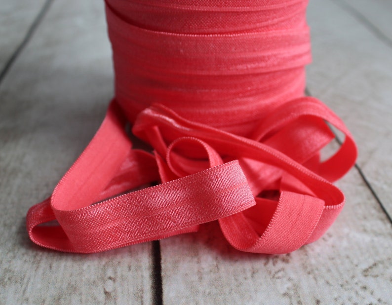 May include: A roll of coral pink elastic fold over elastic, also known as FOE, is shown on a wooden surface. The elastic is soft and stretchy, and it is perfect for sewing projects.