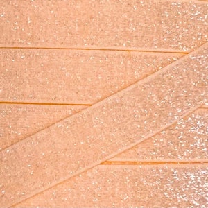 May include: A close-up of peach-colored elastic with a fine silver glitter finish. The elastic is folded and layered, showing the texture and sparkle.