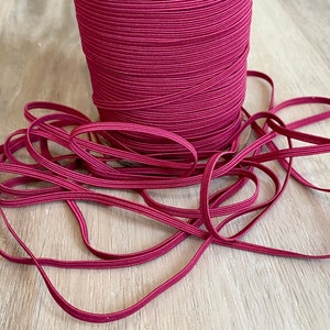 Può includere: Un rotolo di cordoncino elastico bordeaux, con un pezzo di cordoncino disposto su una superficie di legno.