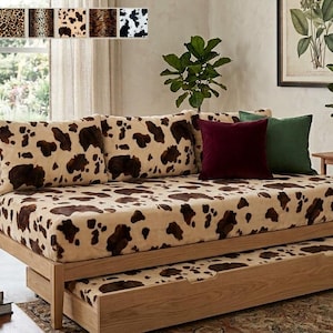 May include: A wooden daybed with a pull-out trundle, both upholstered in a beige fabric with a brown cow-print pattern. The daybed includes two large matching cow-print cushions, one solid burgundy square pillow, and one solid dark green pillow.