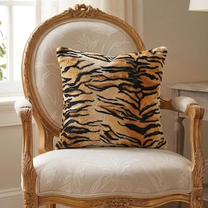 May include: A square throw pillow featuring a realistic tiger stripe pattern in shades of orange, black, and cream. The pillow has a soft, textured surface and is positioned on a light-colored upholstered chair with a carved wooden frame.