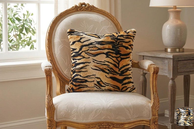 May include: A square throw pillow featuring a realistic tiger stripe pattern in shades of orange, black, and cream. The pillow has a soft, textured surface and is positioned on a light-colored upholstered chair with a carved wooden frame.