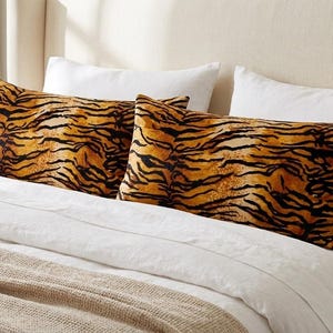 May include: Two rectangular pillowcases featuring a vibrant orange and black tiger stripe pattern. The pillowcases are positioned on a bed with white pillows and a white duvet cover, with a textured beige blanket draped across the bottom of the bed.