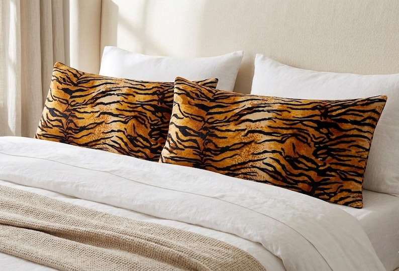 May include: Two rectangular pillowcases featuring a vibrant orange and black tiger stripe pattern. The pillowcases are positioned on a bed with white pillows and a white duvet cover, with a textured beige blanket draped across the bottom of the bed.