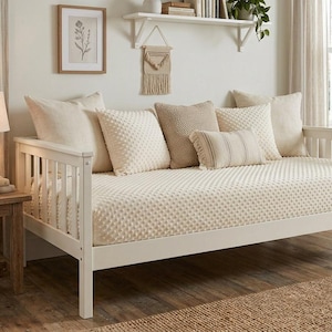 May include: A white wooden daybed frame featuring a slatted back and side panels, topped with a cream-colored textured mattress cover and an assortment of decorative throw pillows in various neutral shades of beige, cream, and light tan.