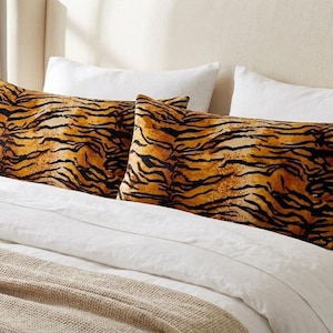 May include: Two rectangular pillowcases featuring a vibrant orange and black tiger stripe pattern. The pillowcases are positioned on a bed with white pillows and a white duvet cover, with a textured beige blanket draped across the bottom of the bed.