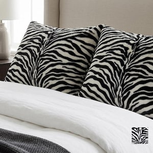 May include: Two square throw pillows featuring a black and white zebra stripe pattern. The pillows are positioned upright on a bed with a white duvet cover and a dark gray textured blanket, with a small square logo visible in the bottom right corner.