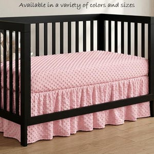 May include: A black wooden crib featuring a pink dimpled minky fabric crib sheet and a matching ruffled crib skirt. The text "Available in a variety of colors and sizes" is printed above the crib. A plush toy and a woven basket are visible.