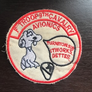 Puede incluir: Un parche circular blanco y rojo con el texto "1st Troop 9th Cavalry Avionics" en un círculo rojo alrededor del borde. Un perro de dibujos animados con un globo de pensamiento que dice "Enciéndelo, funciona mejor" está en el centro del parche.