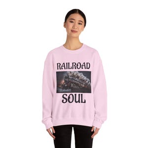 May include: Pale pink sweatshirt featuring the text "RAILROAD SOUL" in black. The central graphic depicts a vintage steam train with white smoke against a dark backdrop. The sweatshirt is a relaxed, casual style.