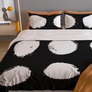 May include: Black and white duvet cover with a large white circle pattern. The duvet cover is on a bed with white pillows and a brown throw blanket.