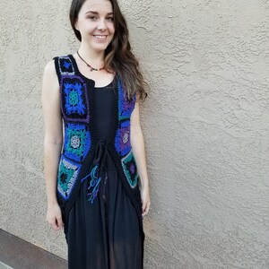 May include: A woman wearing a long black dress with a crocheted vest featuring a colorful geometric pattern. The vest has a tie closure and is made with blue, purple, green, and gray yarn.