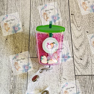 May include: A pink and green glitter filled plastic cup with a straw and fruit inside. The cup is attached to a clear plastic badge holder with a metal clasp. The badge holder has a clear sticker with the text "Glitzy Tiger Co."