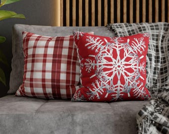 May include: Two decorative throw pillows on a gray couch. One pillow is red with a white snowflake design. The other pillow is red and white plaid.