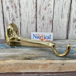 May include: A brass-colored wall hook with a curved design. The hook has a flat base with two screw holes and a curved arm ending in a rounded hook. The text "The nautical place" is visible in the background.