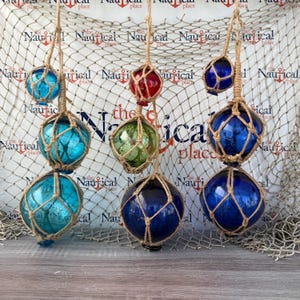 May include: A collection of decorative glass fishing floats in various colors, including blue, green, and red. Each float is encased in a woven jute net and suspended by a jute rope, creating a nautical theme.