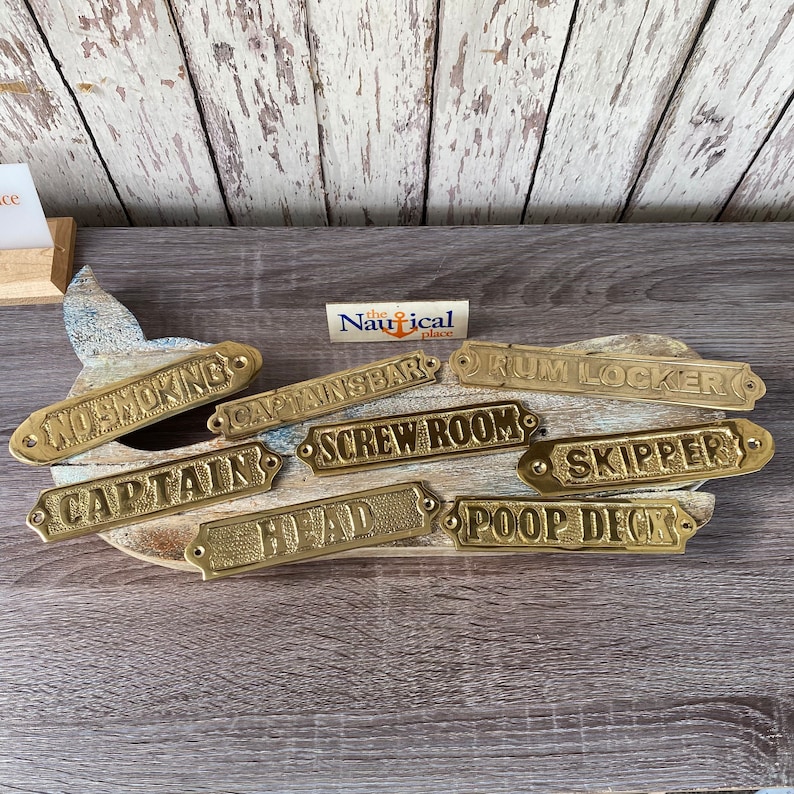 May include: A set of nine gold-coloured metal signs with nautical themed text. The signs are arranged in a semi-circle on a wooden surface. The text on the signs reads: "No Smoking", "Captain's Bar", "Rum Locker", "Screw Room", "Captain", "Head", "Poop Deck", "Skipper", and "The Nautical Place".