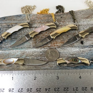 Pu&ograve; includere: Sei piccoli coltelli da tasca in oro con manici in madreperla e lame d'argento sono esposti su una superficie di legno. I coltelli sono attaccati a catene e sono lunghi circa 5 cm. Un righello &egrave; incluso per la scala.
