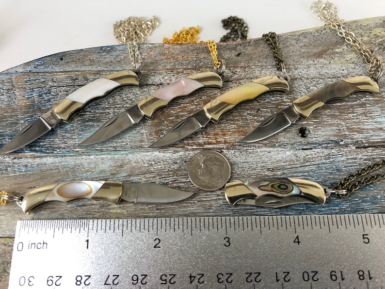 Pu&ograve; includere: Sei piccoli coltelli da tasca in oro con manici in madreperla e lame d'argento sono esposti su una superficie di legno. I coltelli sono attaccati a catene e sono lunghi circa 5 cm. Un righello &egrave; incluso per la scala.