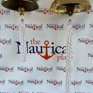 以下が含まれることがあります： 白い背景に「the nautical place」という文字が繰り返し書かれた、白いロープの取っ手がついた金色の船の鐘が2つ。