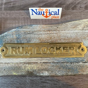 May include: A gold-coloured metal sign with rounded edges and two small holes on either side. The sign reads "RUM LOCKER" in raised letters.