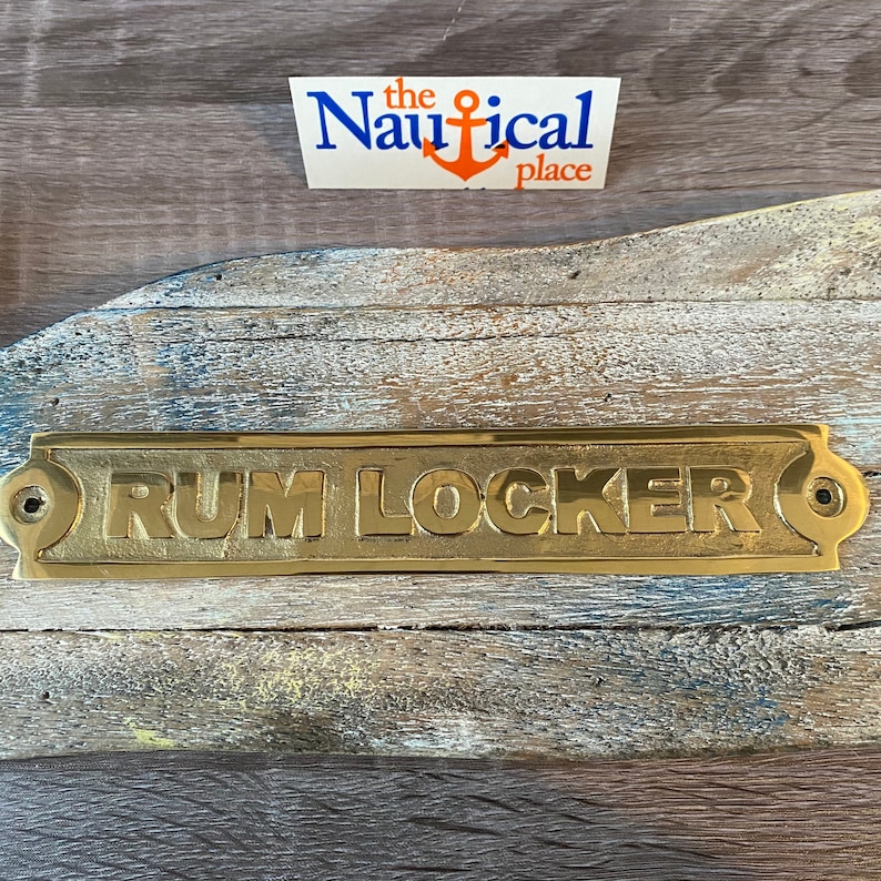 May include: A gold-coloured metal sign with rounded edges and two small holes on either side. The sign reads "RUM LOCKER" in raised letters.