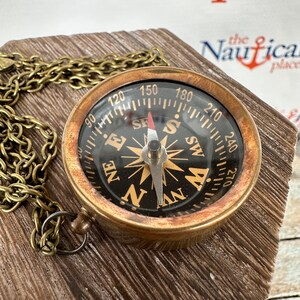 May include: A brass compass necklace with a black face and gold accents. The compass has a chain attached to it and is sitting on a wooden surface. The compass face has the letters N, S, E, and W, as well as numbers from 90 to 270.