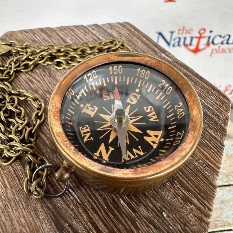 May include: A brass compass necklace with a black face and gold accents. The compass has a chain attached to it and is sitting on a wooden surface. The compass face has the letters N, S, E, and W, as well as numbers from 90 to 270.