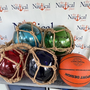 May include: Four glass fishing floats, each encased in a brown rope net, are arranged on a wooden surface. The floats are blue, green, red, and dark blue. A brown basketball is in the foreground.