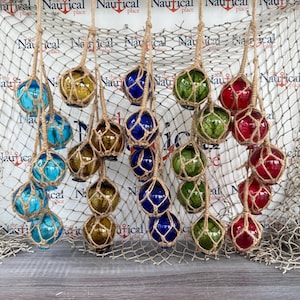 May include: A collection of colorful glass fishing floats in various shades of blue, amber, green, and red, suspended by jute twine. The floats are displayed against a backdrop of a fishing net and a wooden surface.