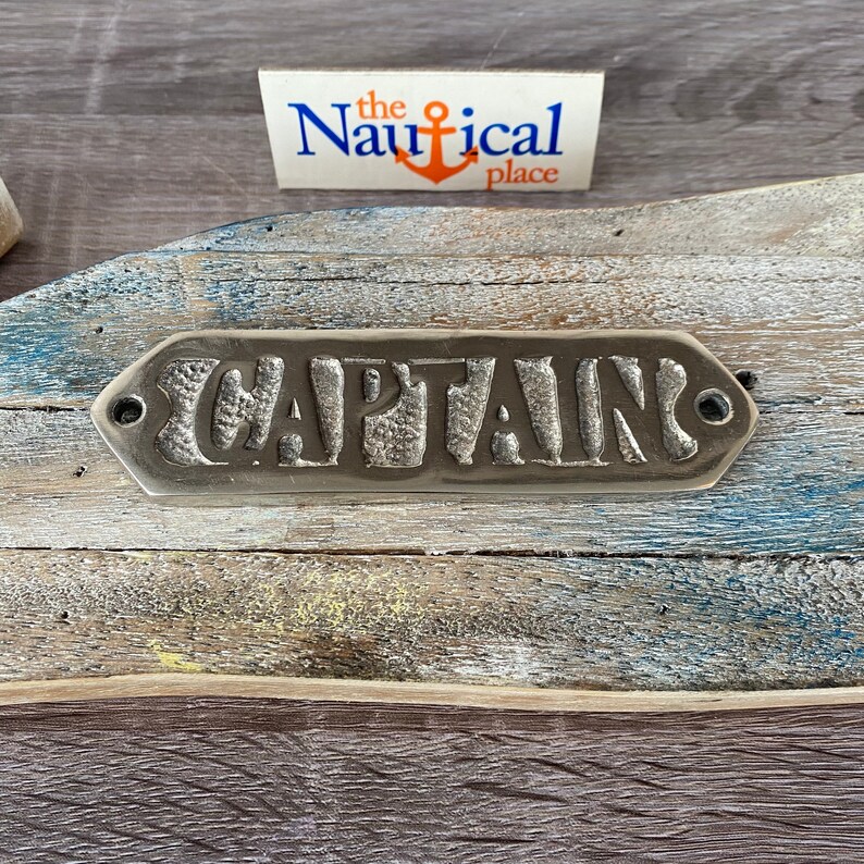 May include: A silver metal sign with the word "CAPTAIN" embossed on it. The sign is mounted on a weathered wooden surface.