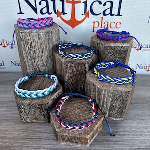 May include: Six different braided bracelets displayed on wooden stands. The bracelets are made of various colors, including blue, red, white, yellow, and purple. The bracelets are all tied with a knot at the end. The stands are all hexagonal in shape and are made of dark wood. The background is a white wall with the words "Nautical place" written in black.