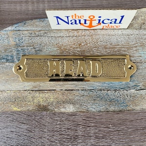 May include: A gold-coloured brass sign with the word "HEAD" embossed on it. The sign has a textured surface and rounded edges. It is mounted on a wooden surface.